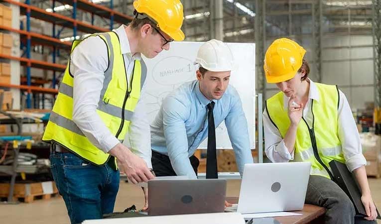 Two men and a woman wear hard hats in warehouse area, looking at laptops as they implement ERP software.