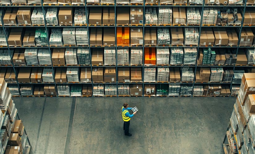 Man with tablet stands in warehouse aisle using software for inventory management.