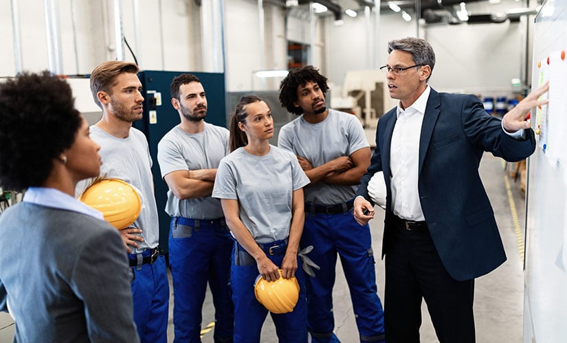 Five factory workers stand around a man pointing to a board representing the importance of scalable QMS and ERP software for growing operations.
