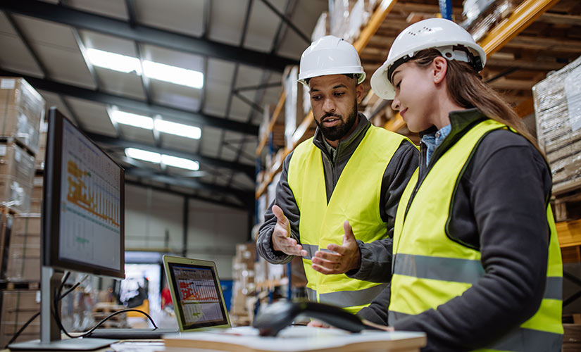 A man and woman in hardhats stand before laptop and barcode scanner in warehouse environment using ERP software.