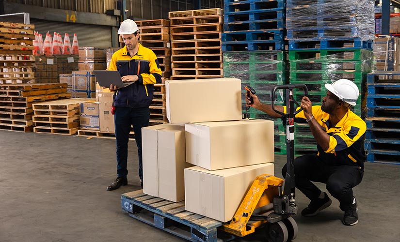 A man stands in warehouse in front of pallet of boxes, while another crouches as he uses a barcode scanner as part of closed-loop manufacturing traceability.