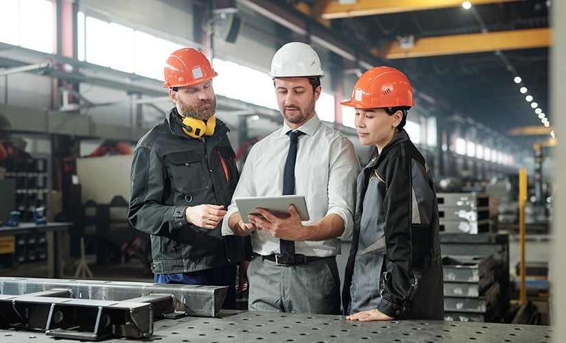 Two men and a woman wear hardhats in factory. All stand looking at laptop as they use ERP software to track manufacturing rework and costs.