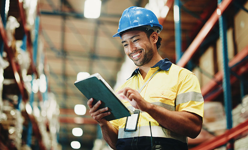 Man in blue hardhat stands in warehouse using a digital tablet to manage supplier quality.