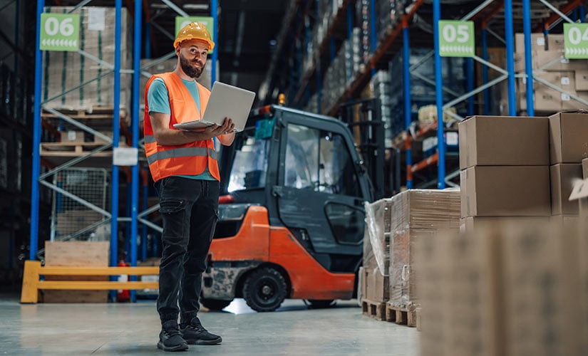 Warehouse worker using laptop in front of shelves. Returns management software like QT9 ERP automates returns processing, helping manufacturers eliminate costly delays and maintain compliance.