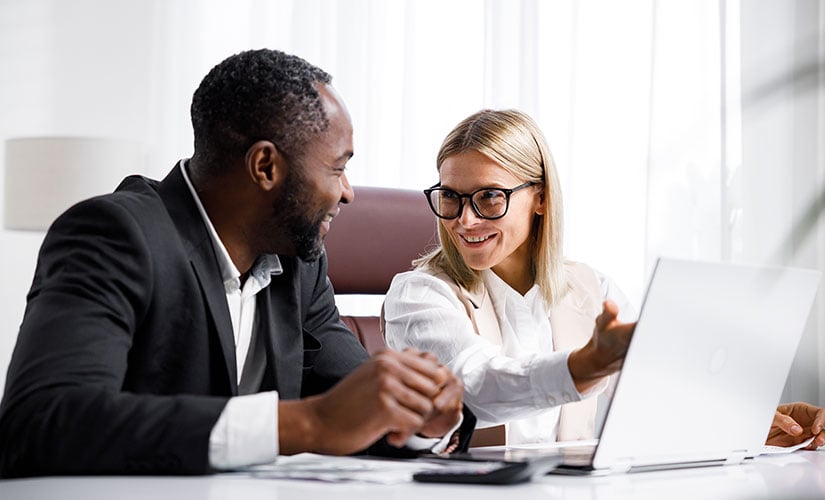 A man and woman sit next to each other at a desk, while the woman gestures to a laptop screen as they discuss regulatory compliance software.
