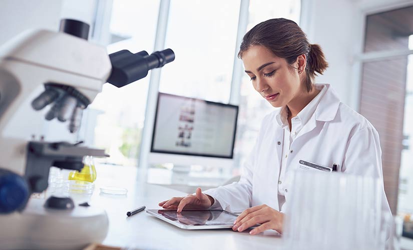 Woman in a lab coat sits at desk with microscope in front and computer screen in back, looking at a computer tablet and learning how to avoid document control audit findings.