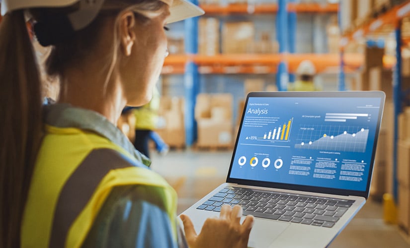 Woman in hard hat stands in warehouse looking at laptop screen that displays ERP forecasting software.