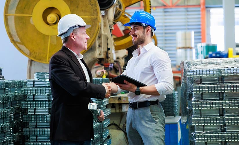 A man in a suit and hardhat shakes the hand of another man in a hardhat holding a laptop. Contract manufacturing is managed seamlessly within QT9 ERP.