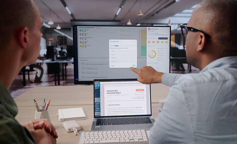 Two men sit in front of a laptop, keyboard and computer screen, managing supplier risk through integrated QMS and ERP software.
