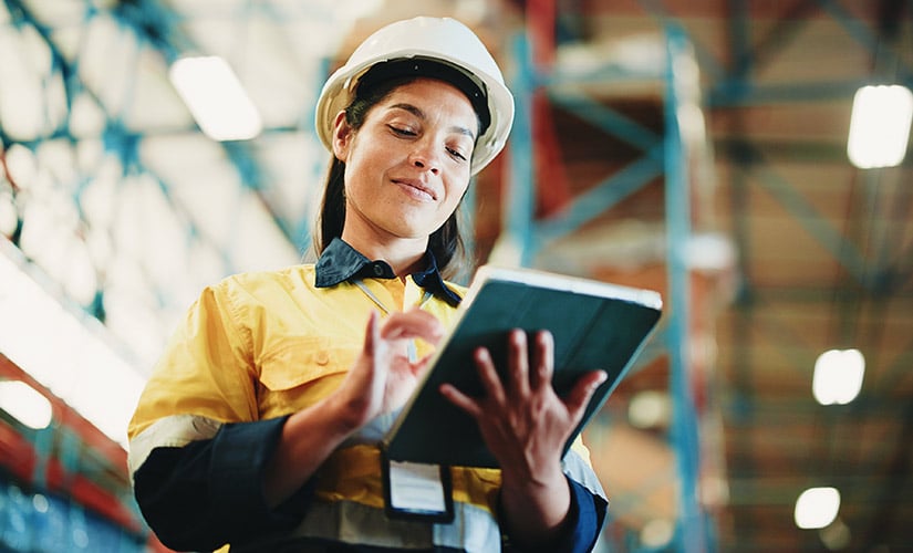 Woman in hardhat in warehouse using a tablet and ERP + QMS software to manage supply chain traceability.