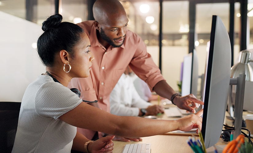 A seated woman and standing man point to a computer screen as they look at data in their employee training management software..