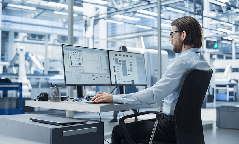 Man in manufacturing facility sits in front of two computer screens managing ERP implementation.