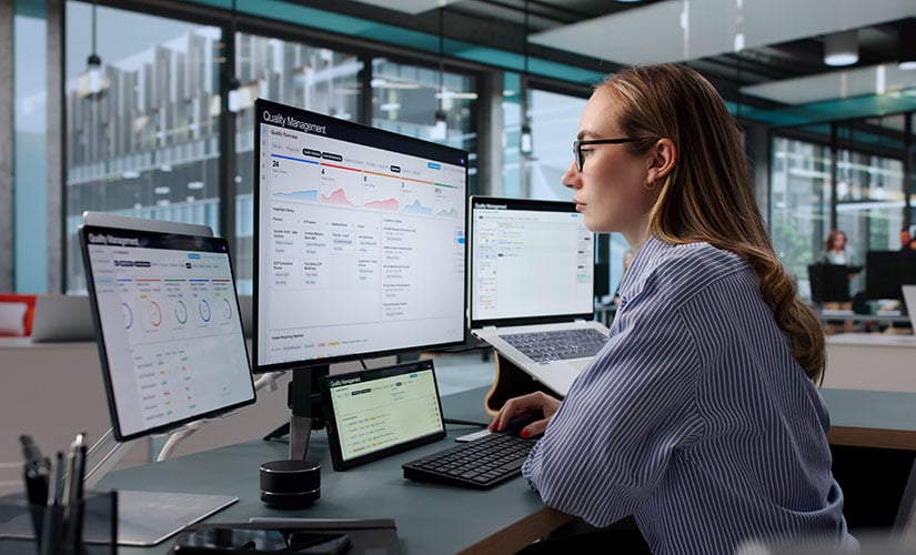 Woman sits in front of two computer screens displaying quality management software, undertaking a supplier risk assessment.